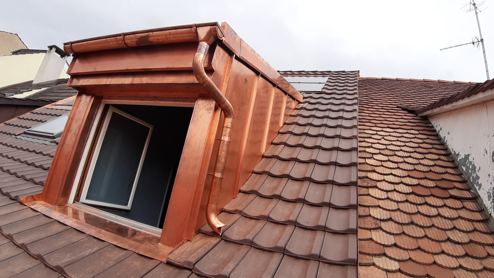 Miedziane rynny i obróbki blacharskie wokół okna dachowego, widok na dach z brązową dachówką ceramiczną. Nowe orynnowanie.