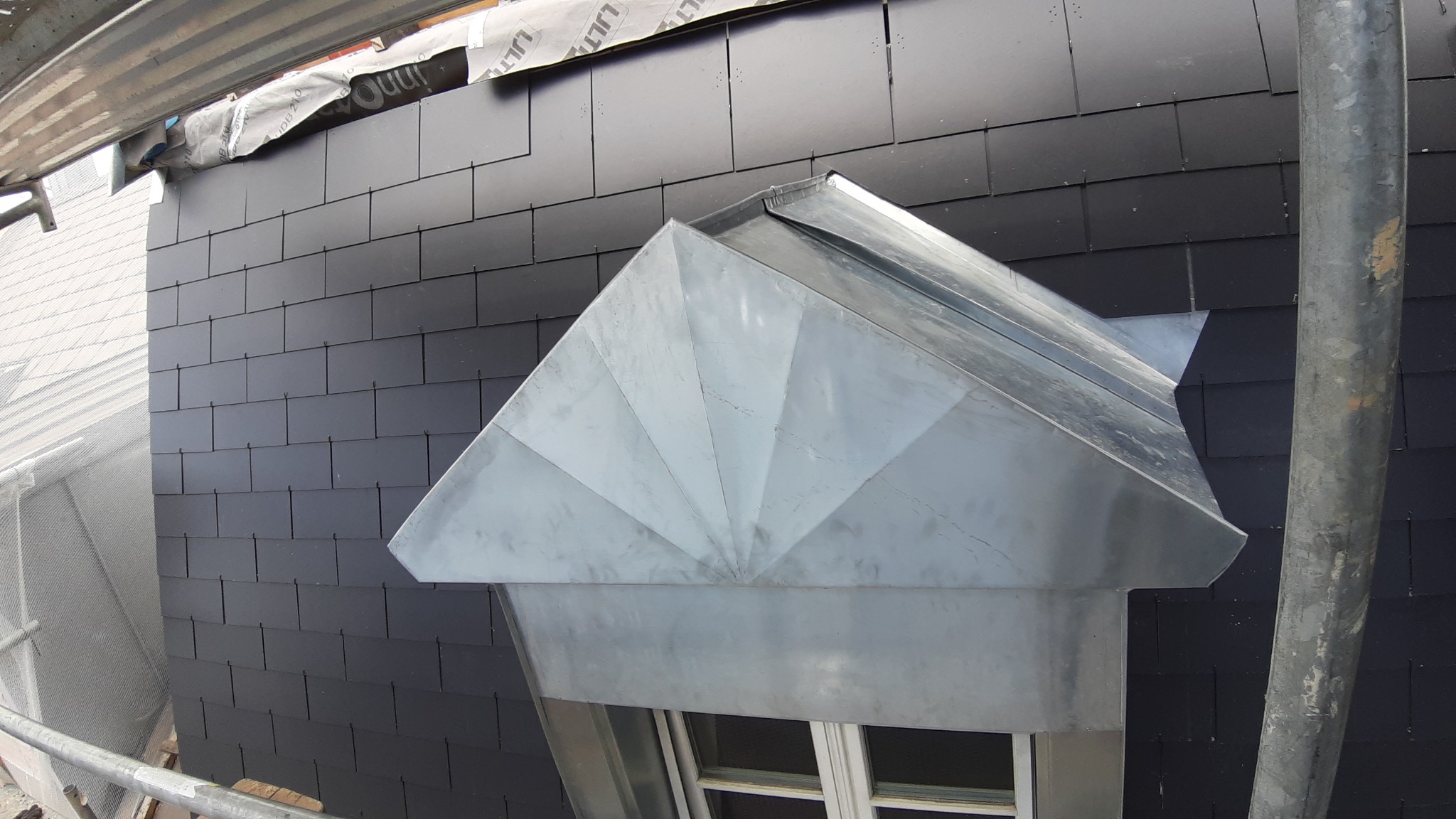 Szczegółowe ujęcie okna dachowego z metalową obudową w kształcie wachlarza na tle czarnego dachu z łupka.