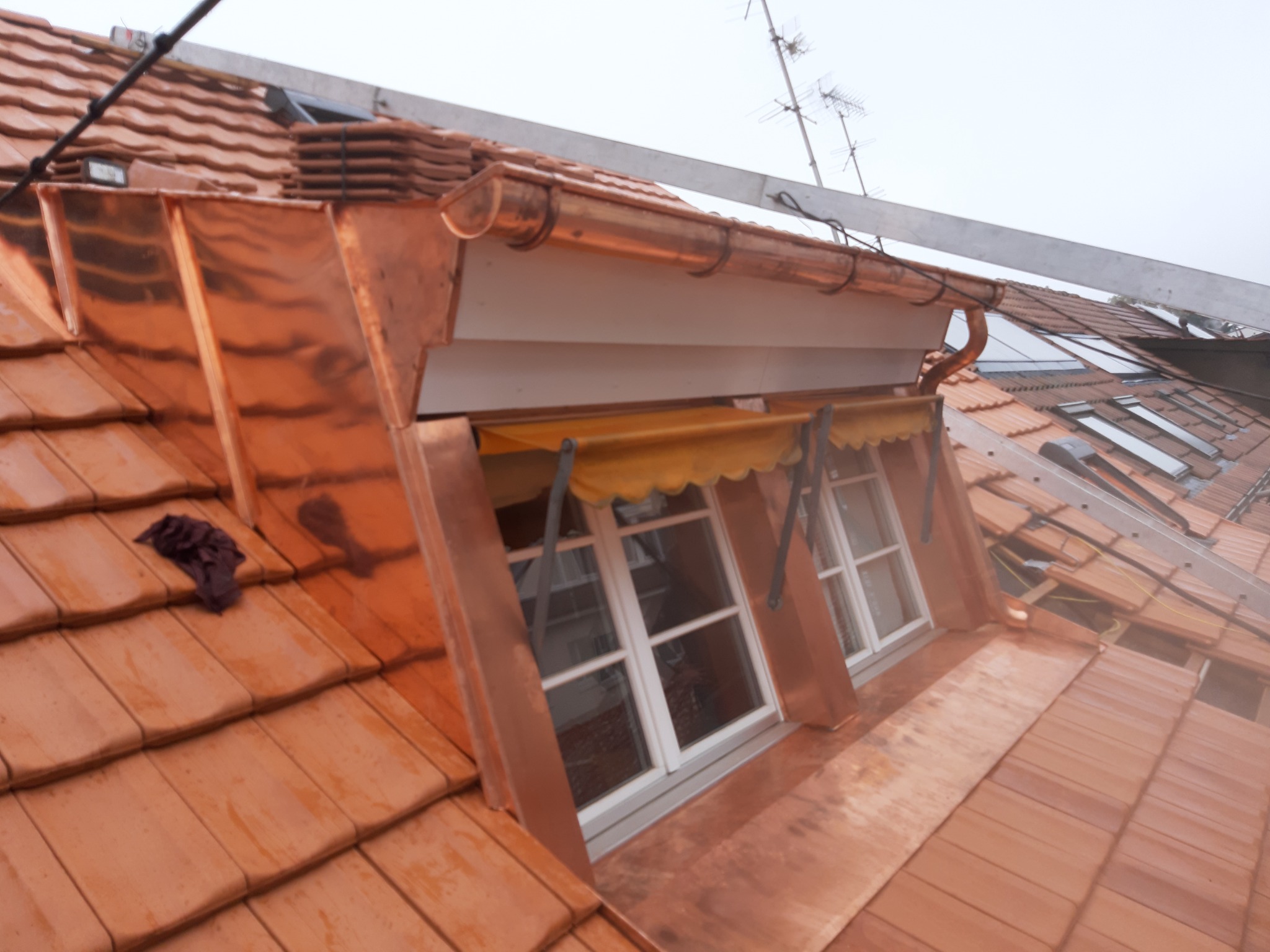 Miedziane obróbki blacharskie okna dachowego na dachu z dachówki ceramicznej, widoczna drabina i antena.