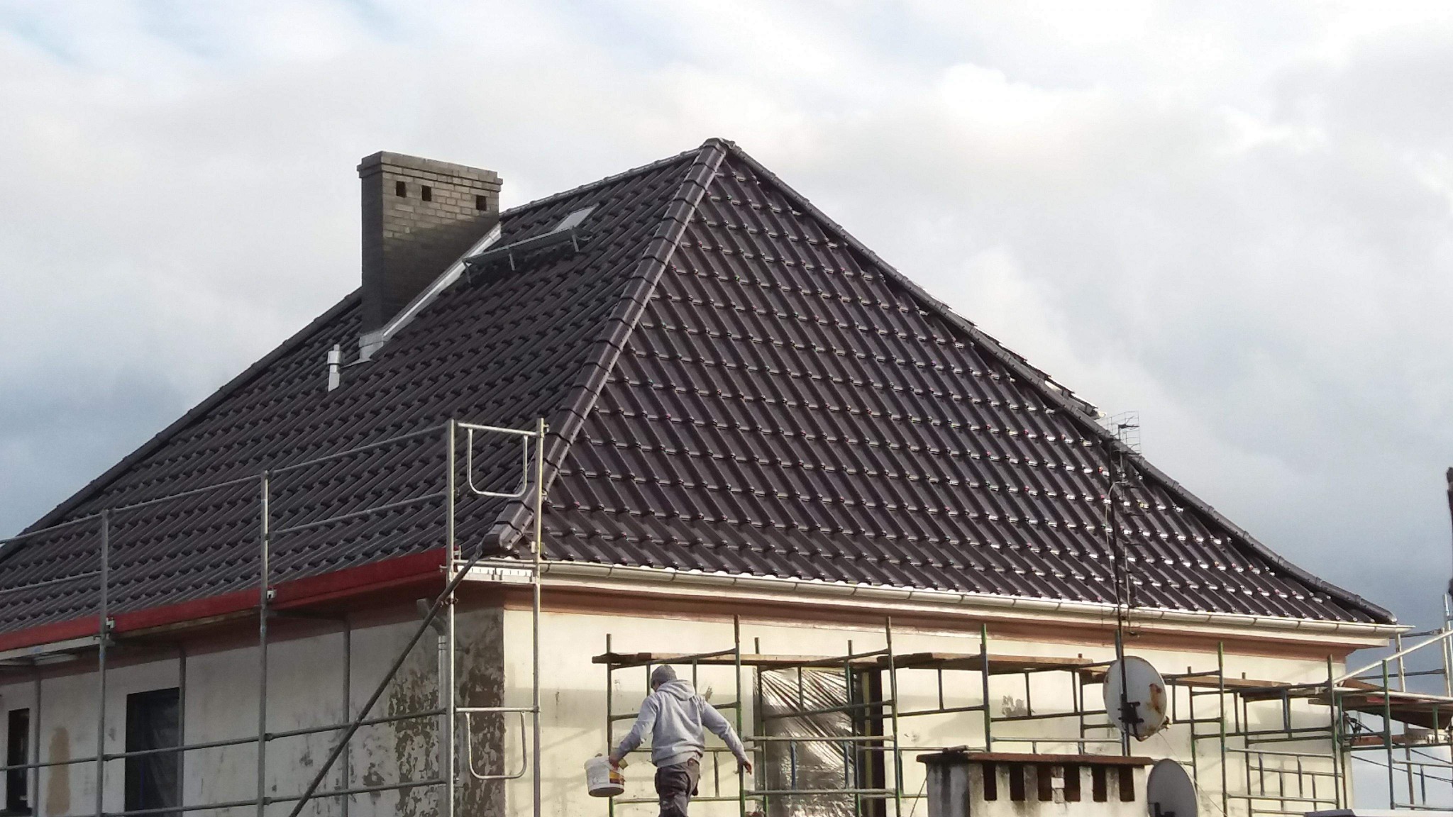 Pracownik na rusztowaniu z wiadrem farby, na tle domu z nowym, brązowym dachem z dachówki ceramicznej i ceglanym kominem.