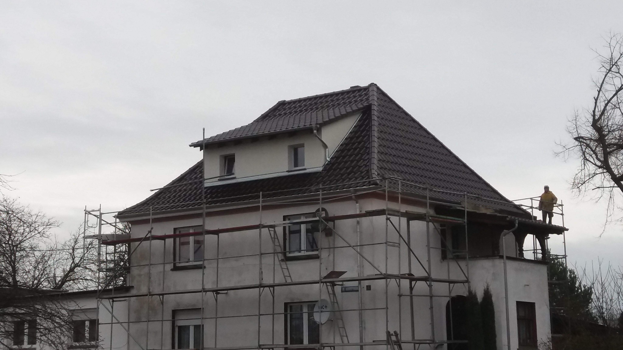Dom jednorodzinny w trakcie renowacji dachu, z widocznymi rusztowaniami i pracownikiem w żółtej kurtce na wysokości, na tle pochmurnego nieba.