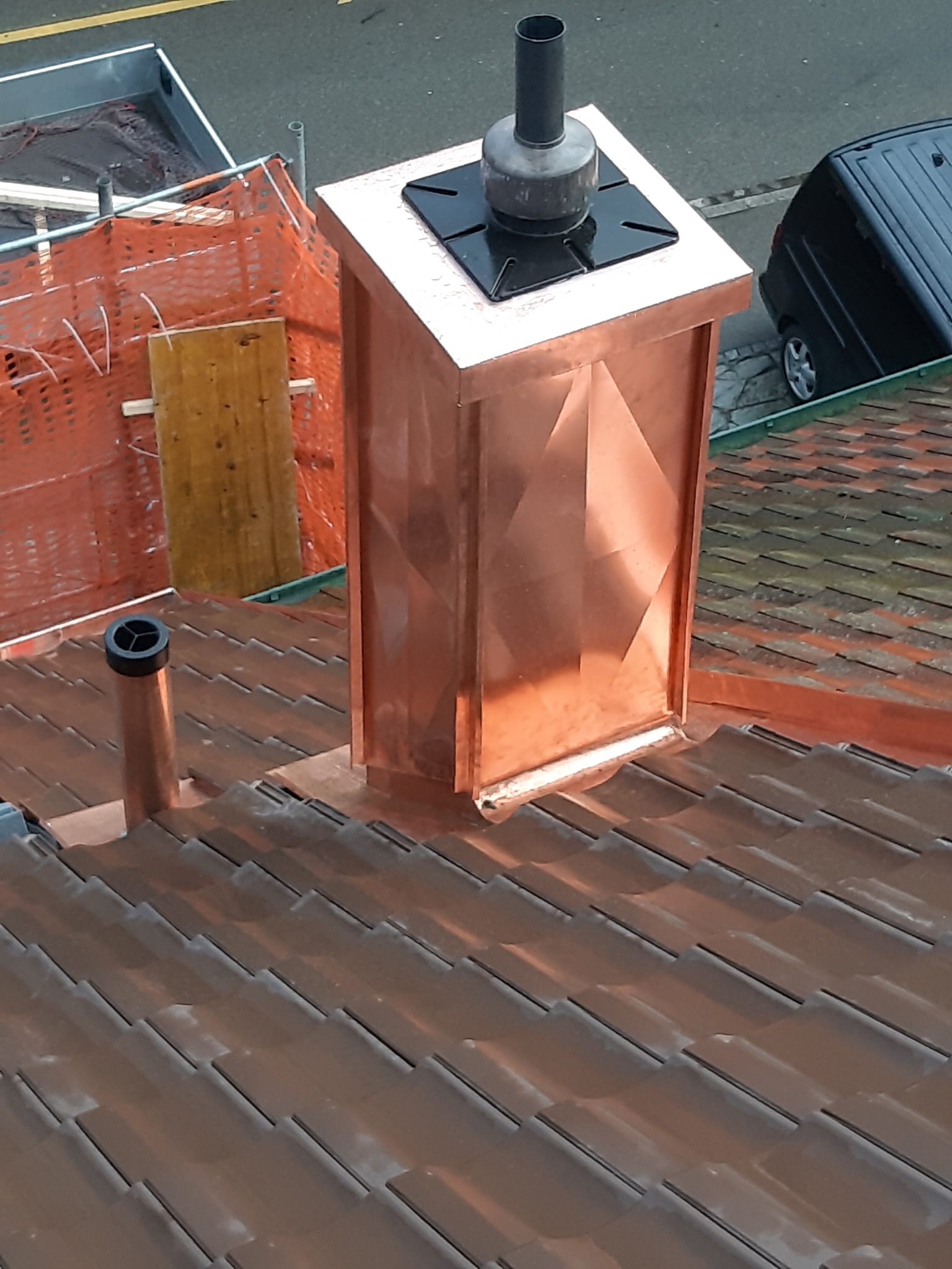Miedziany komin z dekoracyjnymi panelami na dachu z brązowej dachówki ceramicznej, widoczny fragment rusztowania i czarny samochód w tle.