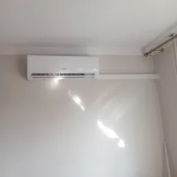 Zamontowany biały klimatyzator na ścianie w jasnym pomieszczeniu, widoczne rury instalacyjne, fragment szarej kanapy w dolnej części kadru.