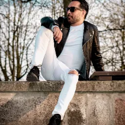 Mężczyzna w okularach przeciwsłonecznych, białych spodniach z dziurą na kolanie i czarnej skórzanej kurtce siedzi na betonowym murku na tle drzew bez liści.