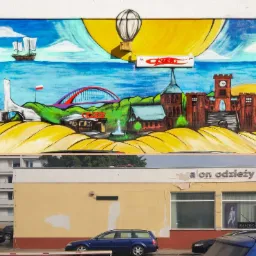 Mural na ścianie budynku przedstawiający krajobraz morski z latarnią, statkiem, mostem, zamkiem i balonem, poniżej widoczny budynek sklepu odzieżowego i zaparkowane samochody.