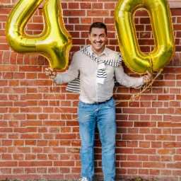 Uśmiechnięty mężczyzna w jeansach i białej koszuli, stojący na tle ceglanej ściany, trzymający złote balony w kształcie cyfr '4' i '0'.