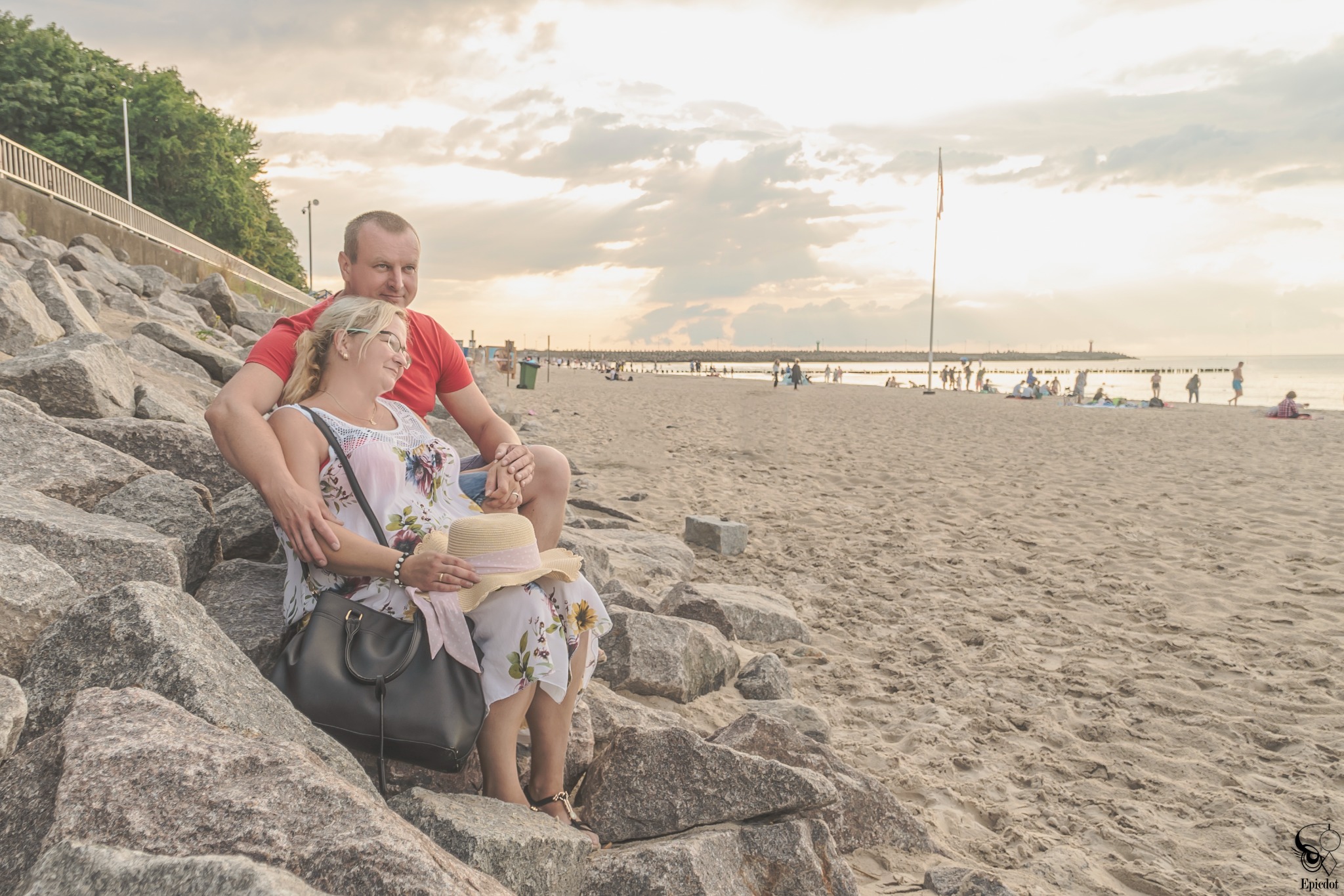 Para w średnim wieku siedzi na kamieniach na plaży w Kołobrzegu podczas sesji ciążowej, w tle morze i zachodzące słońce.
