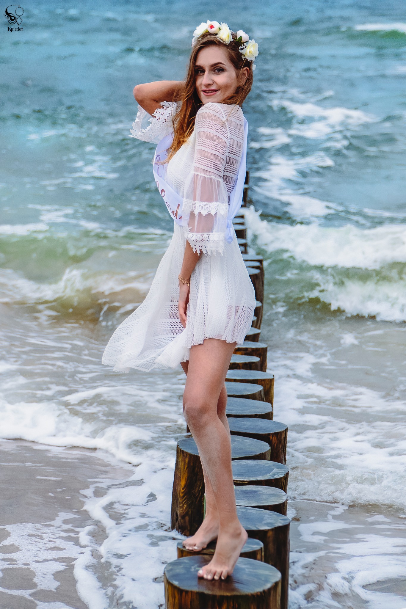 Uśmiechnięta kobieta w białej koronkowej sukience i wianku na głowie, stojąca na drewnianych palach wchodzących w morze, z szarfą przewieszoną przez ramię