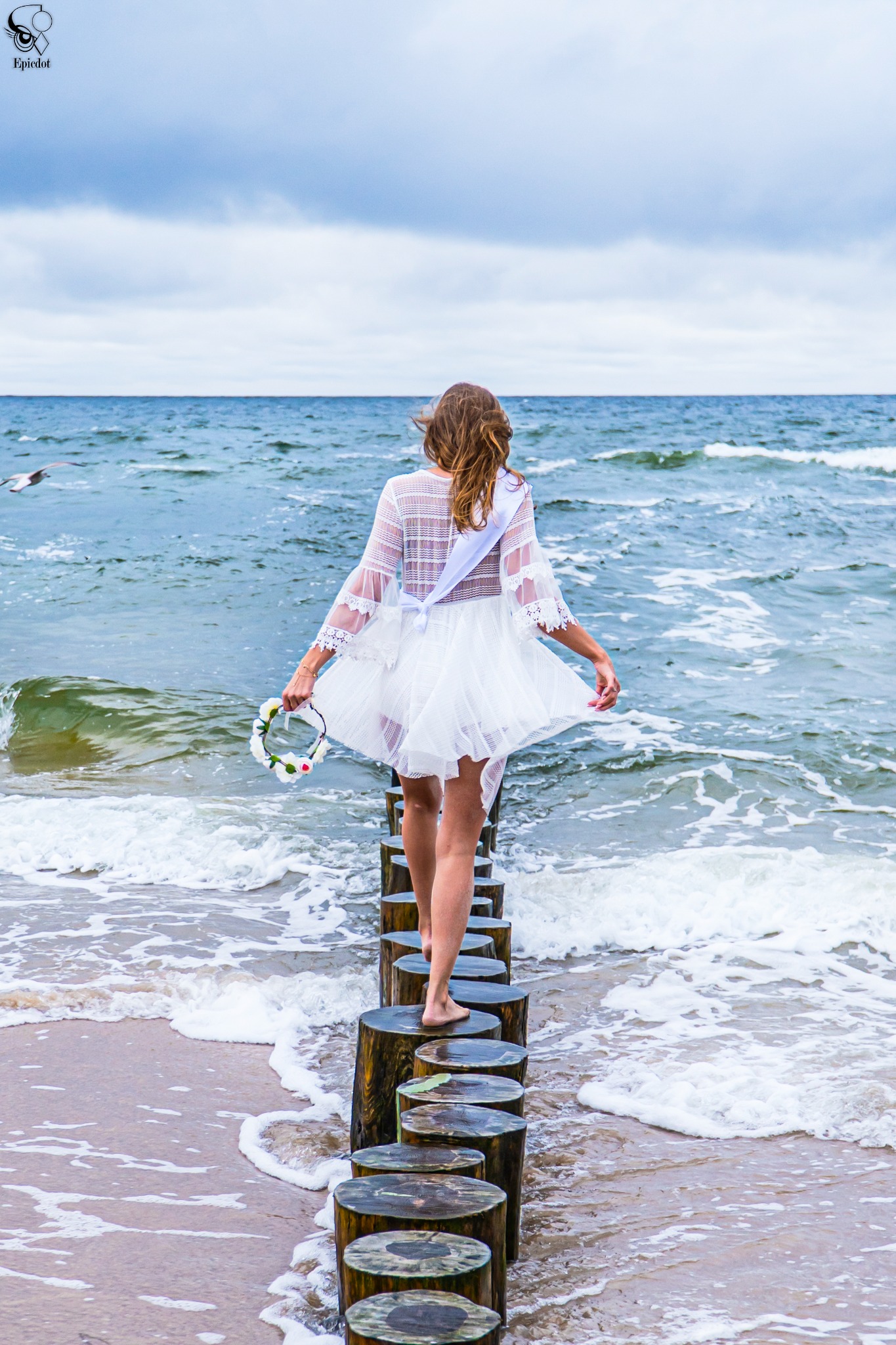Kobieta w białej sukience i z wiankiem w ręku idzie po drewnianych palach w morzu, widok z tyłu, w tle wzburzone fale i pochmurne niebo.