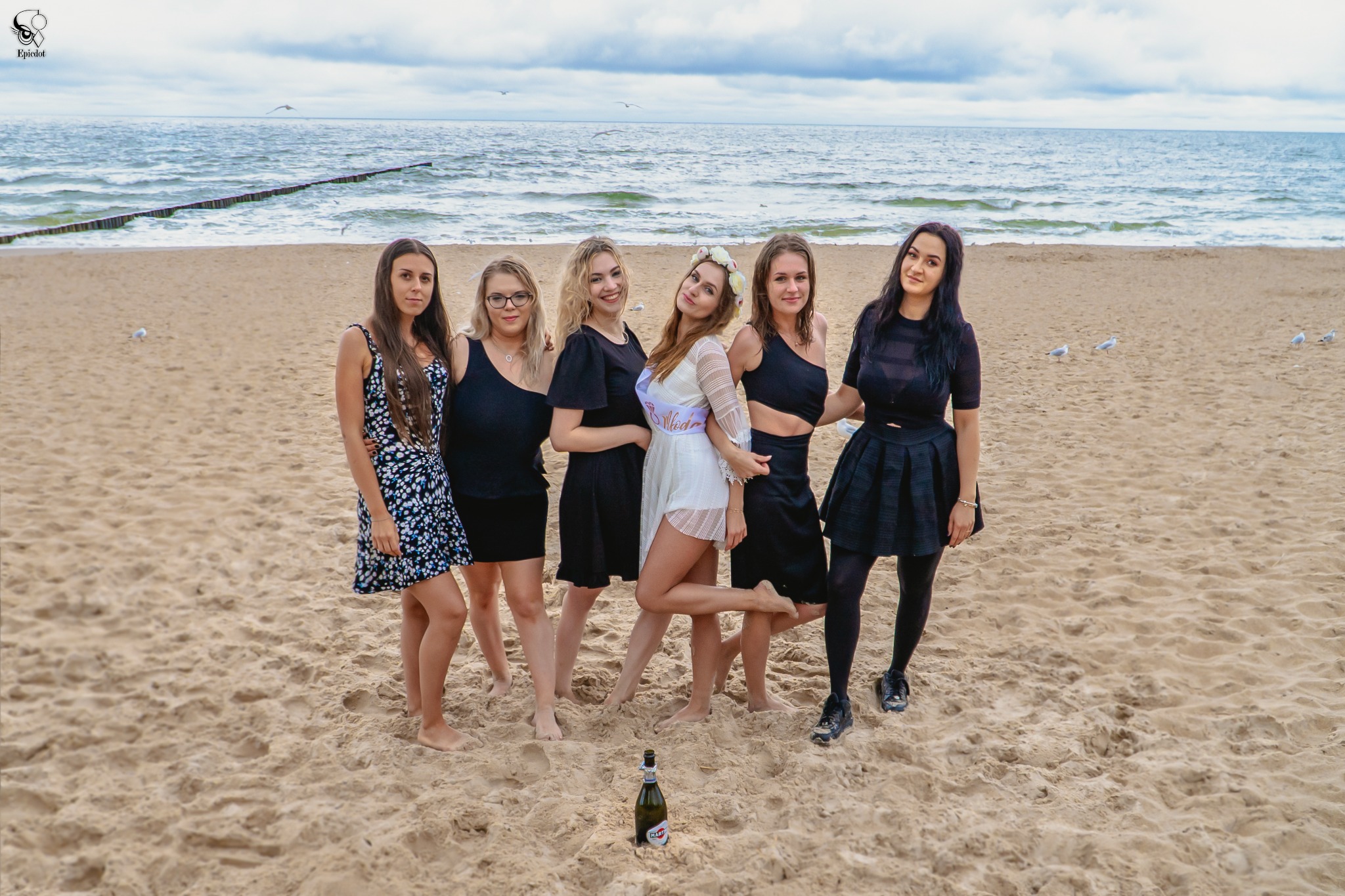 Sześć młodych kobiet na piaszczystej plaży, jedna z szarfą 'Wieczór Panieński', w tle morze i mewy, na piasku butelka Martini.
