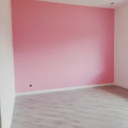 Pomalowany na różowo pokój z panelami podłogowymi w odcieniach szarości, widoczne białe listwy przypodłogowe i oświetlenie sufitowe.