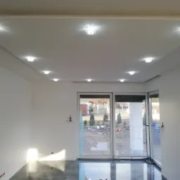 Jasne, przestronne wnętrze po remoncie, z błyszczącą szarą podłogą z płytek, białymi ścianami z widocznymi puszkami elektrycznymi i sufit podwieszany z wbudowanymi kryształowymi lampami. Przez duże...