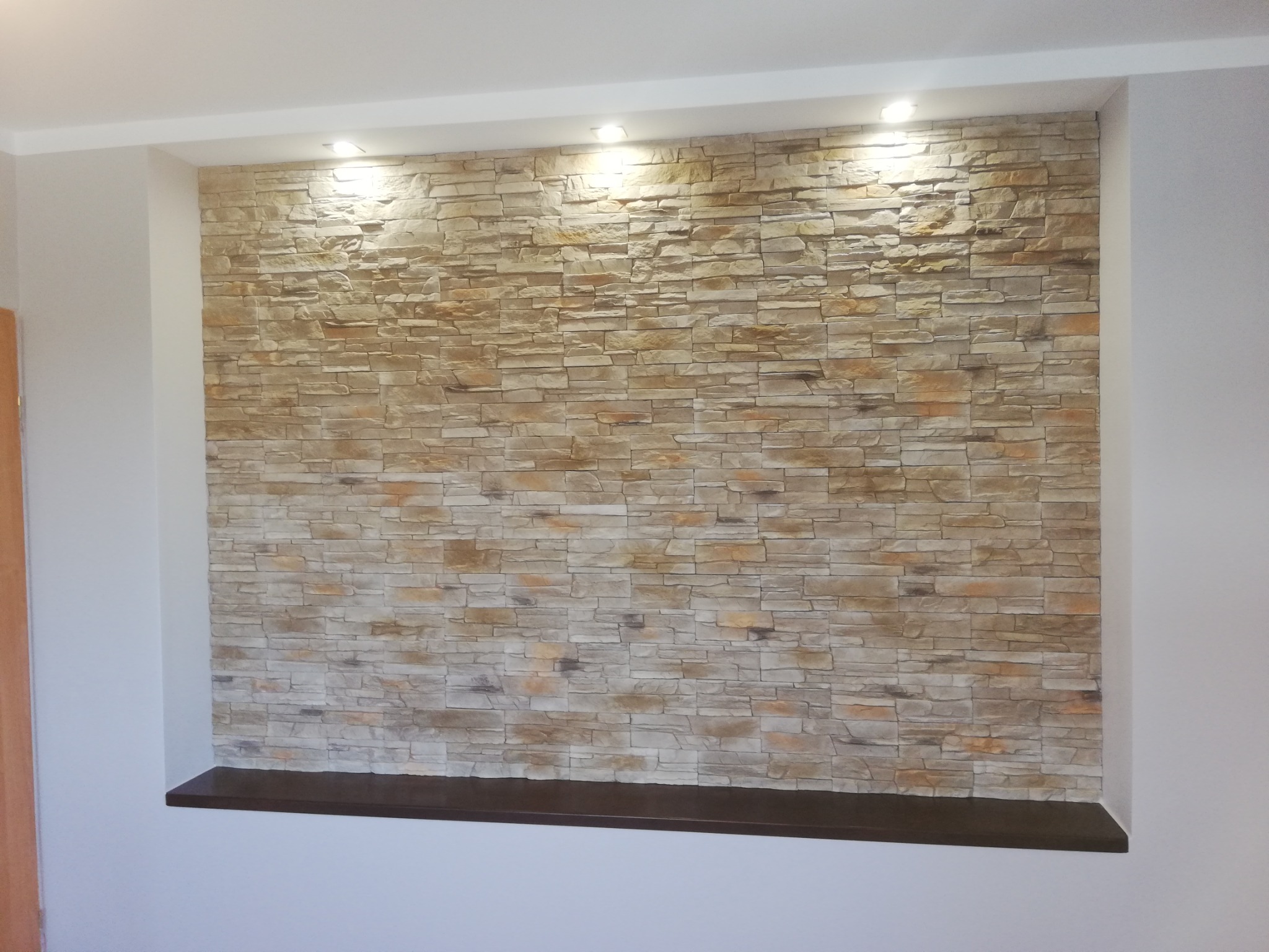 Ściana akcentowa z dekoracyjnej cegły w odcieniach beżu i brązu, podświetlona trzema punktowymi lampami sufitowymi, z ciemnobrązową półką u dołu.