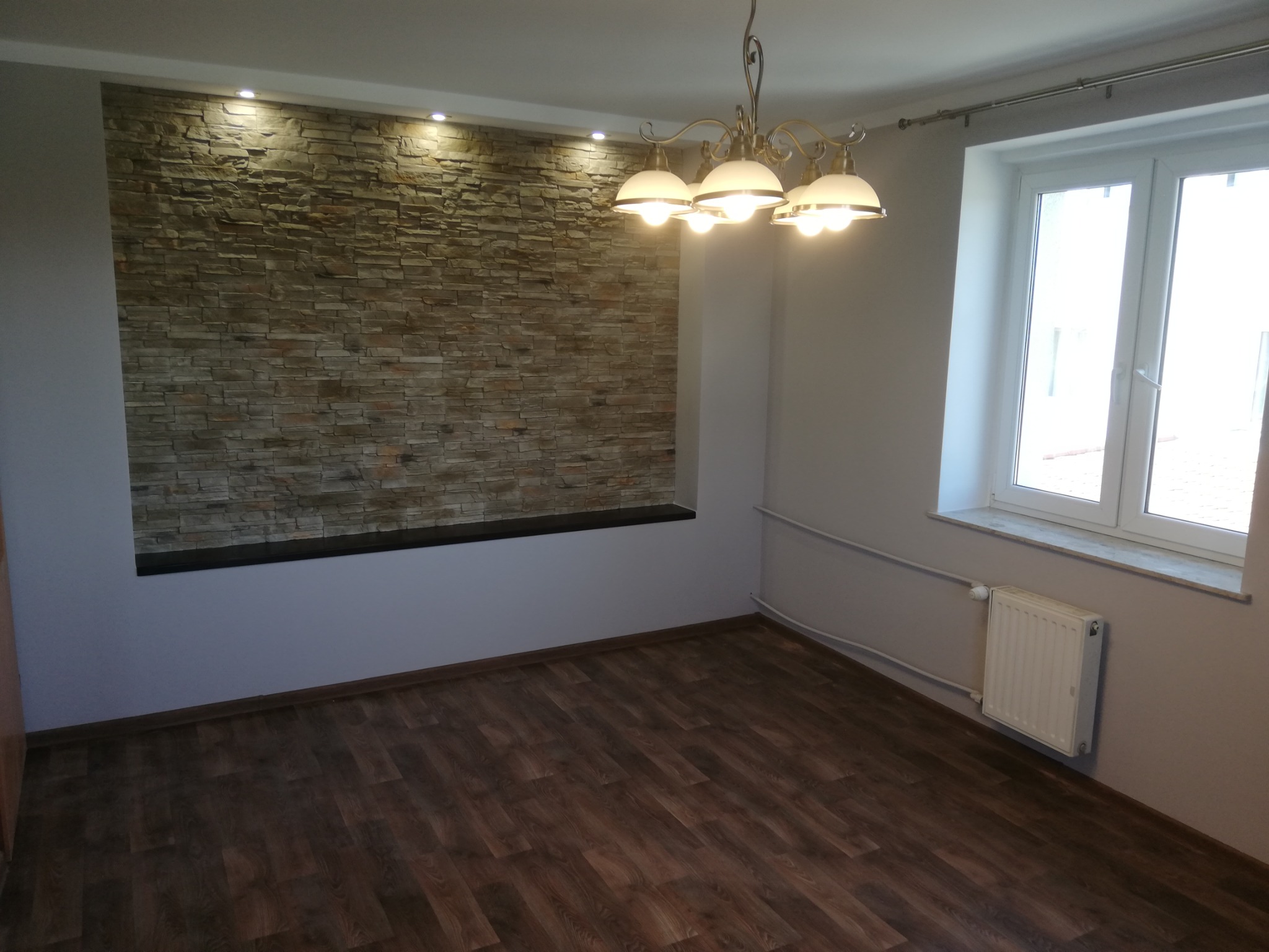 Wykończone wnętrze pokoju z dekoracyjną ścianą z kamienia, oświetloną punktowymi lampami, drewnianą podłogą, białym oknem i grzejnikiem, oświetlone przez wiszącą lampę z kilkoma kloszami.