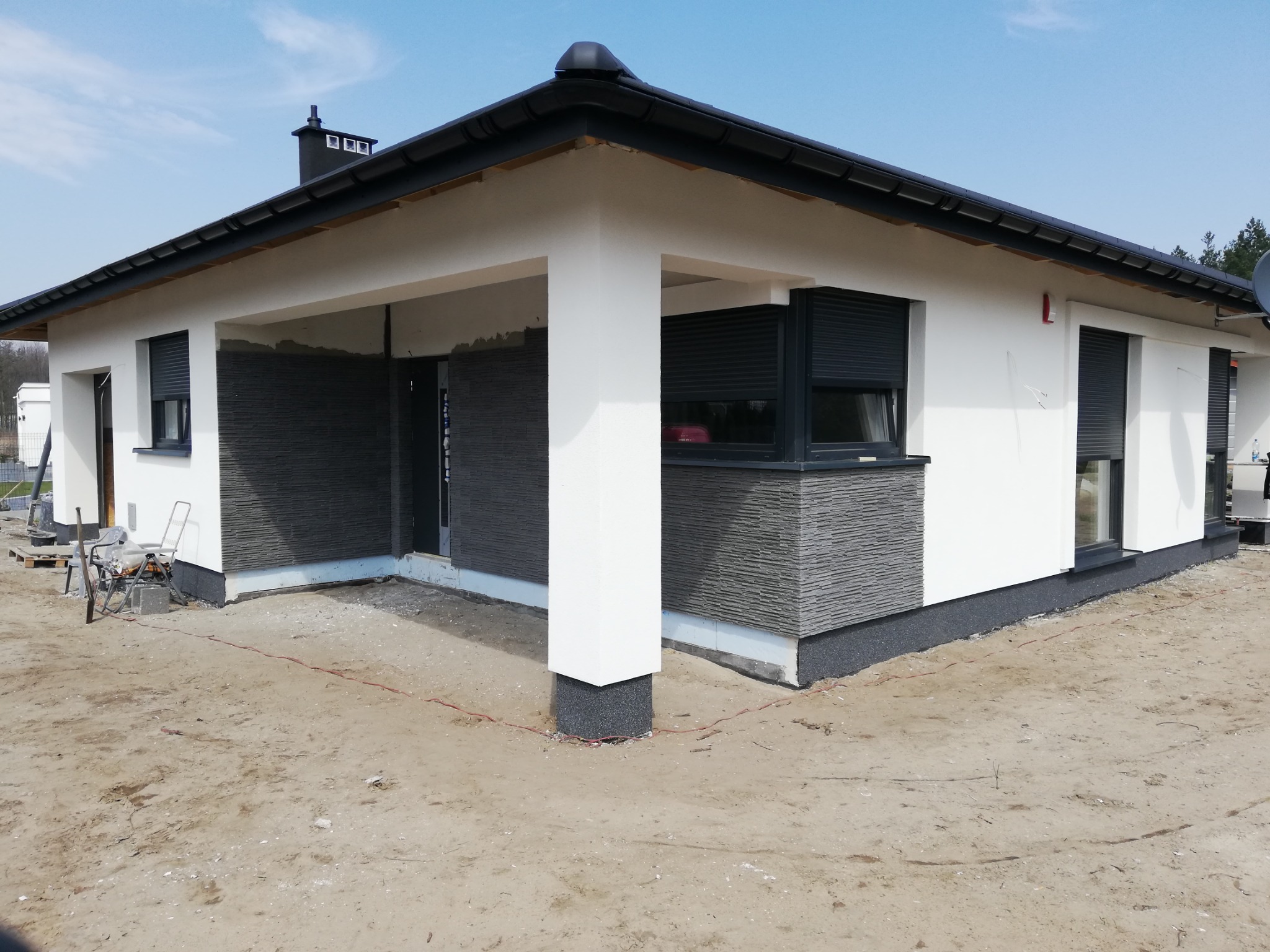 Nowoczesny dom jednorodzinny z białymi ścianami, szarą elewacją z kamienia i czarnymi roletami zewnętrznymi, widok z zewnątrz na etapie wykończenia terenu.