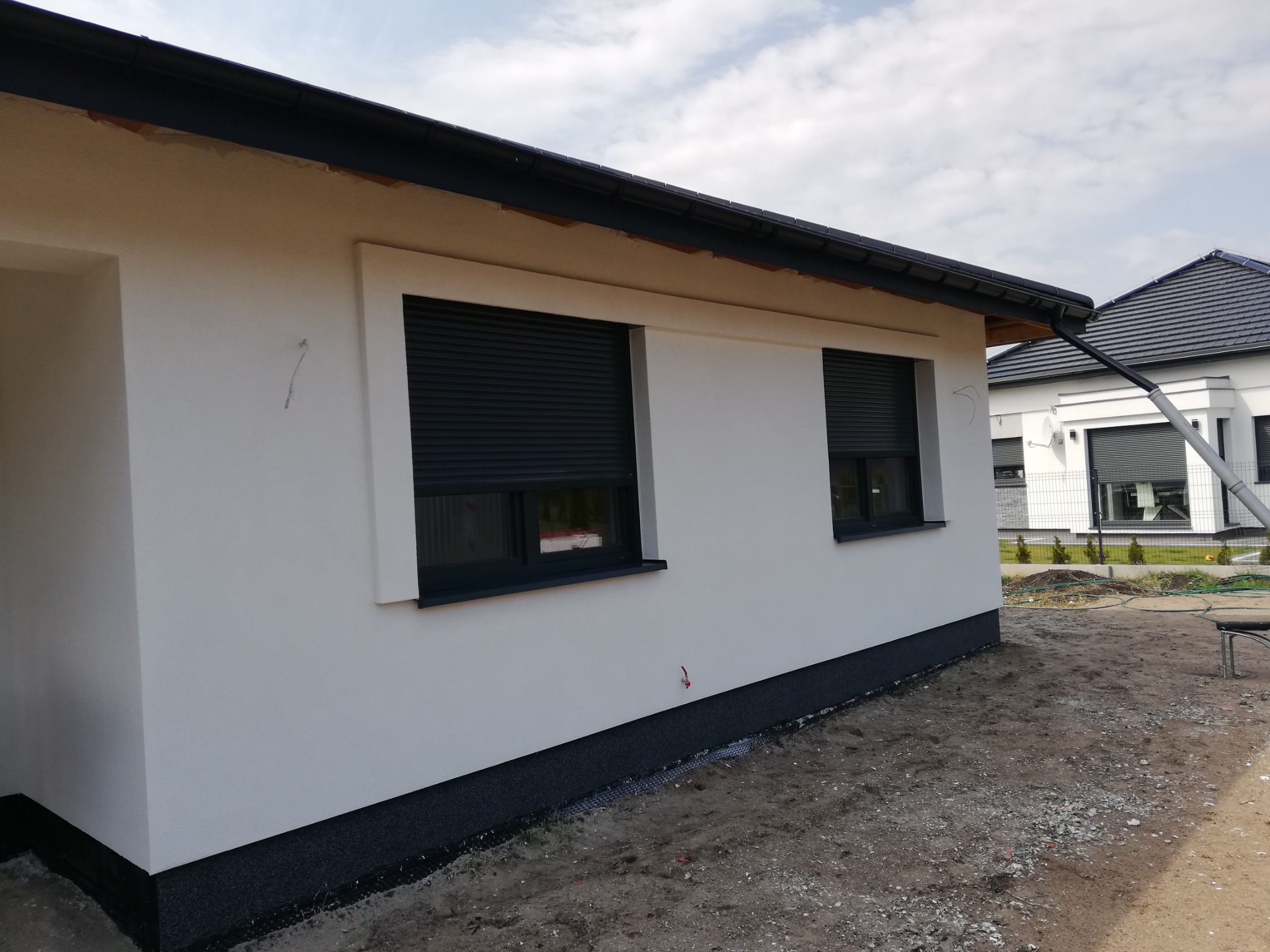 Nowoczesny, parterowy dom z białymi ścianami, czarną podbitką dachową i opuszczonymi roletami zewnętrznymi w kolorze antracytowym, widok z zewnątrz w pochmurny dzień.