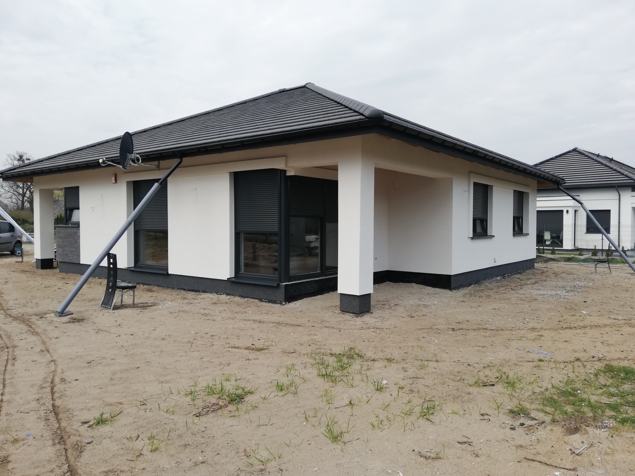Nowoczesny, parterowy dom z białymi ścianami i grafitowym dachem, z zamontowanymi roletami zewnętrznymi i anteną satelitarną, widoczny w surowym otoczeniu placu budowy z piaskiem i prowizorycznym...