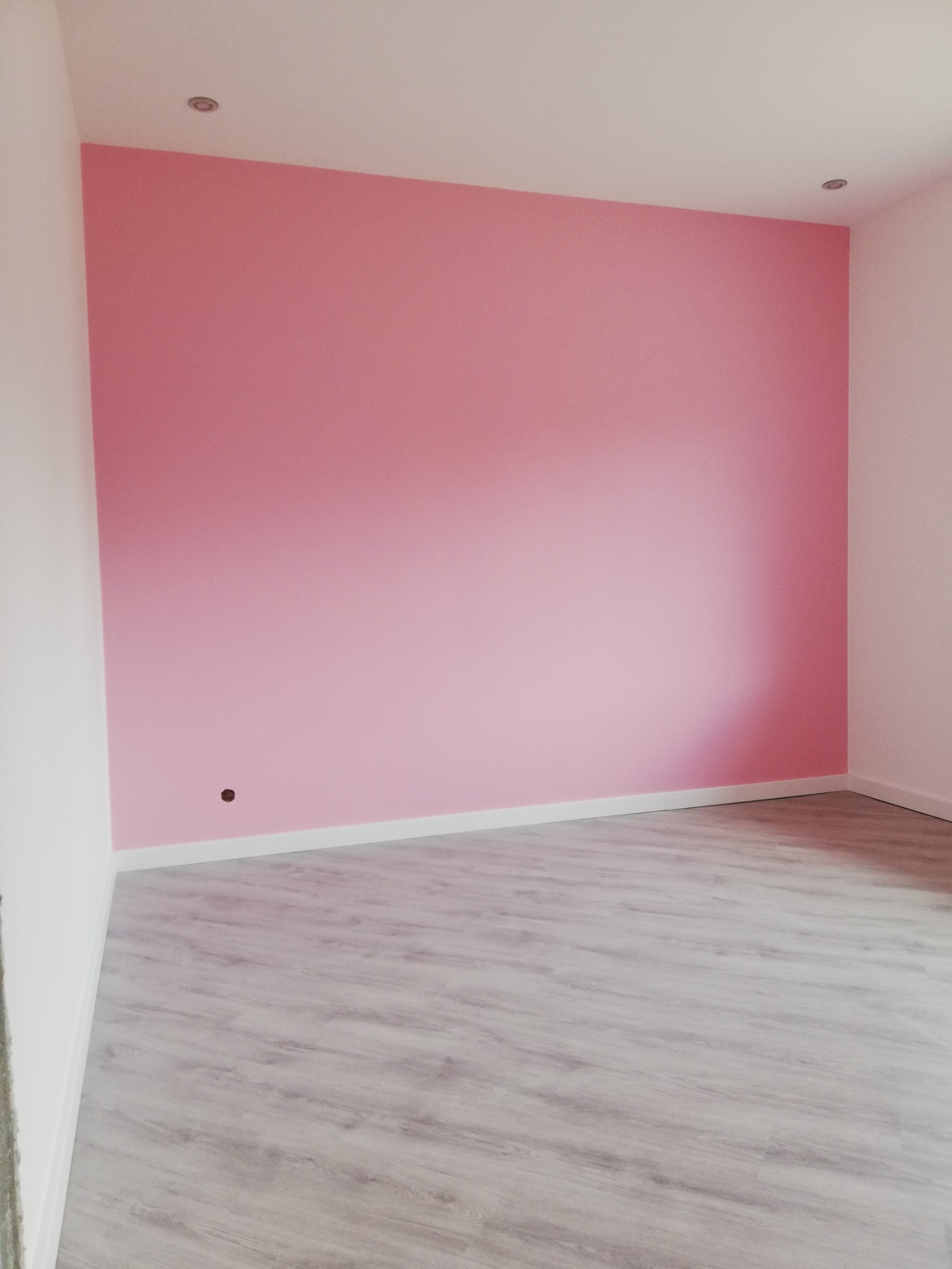 Pomalowany na różowo pokój z panelami podłogowymi w odcieniach szarości, widoczne białe listwy przypodłogowe i oświetlenie sufitowe.