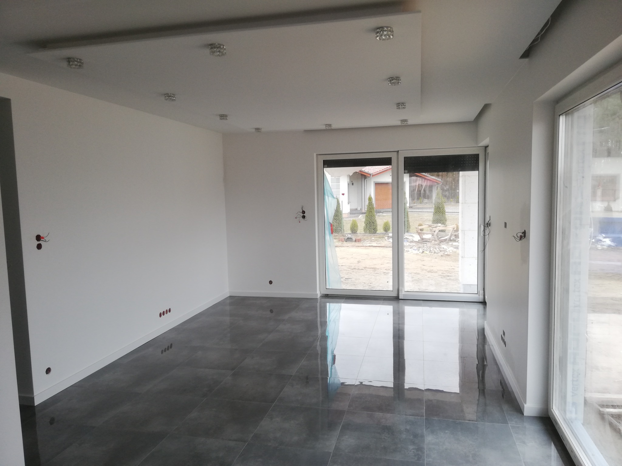 Nowoczesne wnętrze z szarą, błyszczącą podłogą z płytek, białymi ścianami z przygotowanymi punktami elektrycznymi i oknami przesuwnymi z widokiem na ogród w trakcie aranżacji.