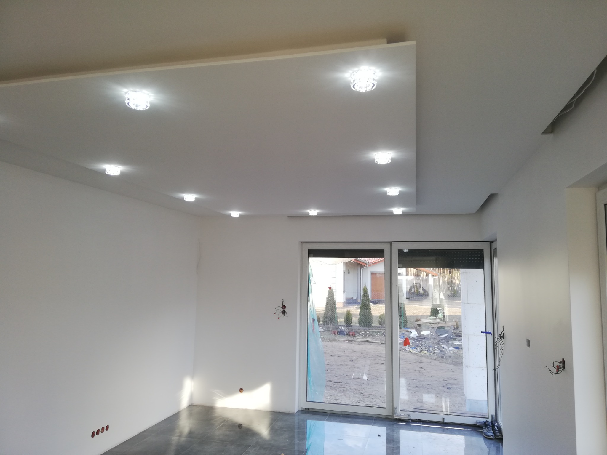 Nowoczesne wnętrze z podwieszanym sufitem z punktowym oświetleniem LED, szara, błyszcząca podłoga, duże okno tarasowe z widokiem na ogród w trakcie prac wykończeniowych.