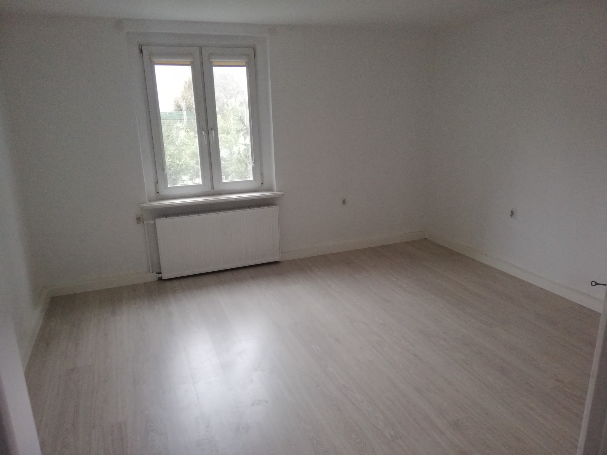 Pusty pokój z panelami podłogowymi w odcieniach szarości, białe ściany, okno z widokiem na zieleń i grzejnik pod oknem.