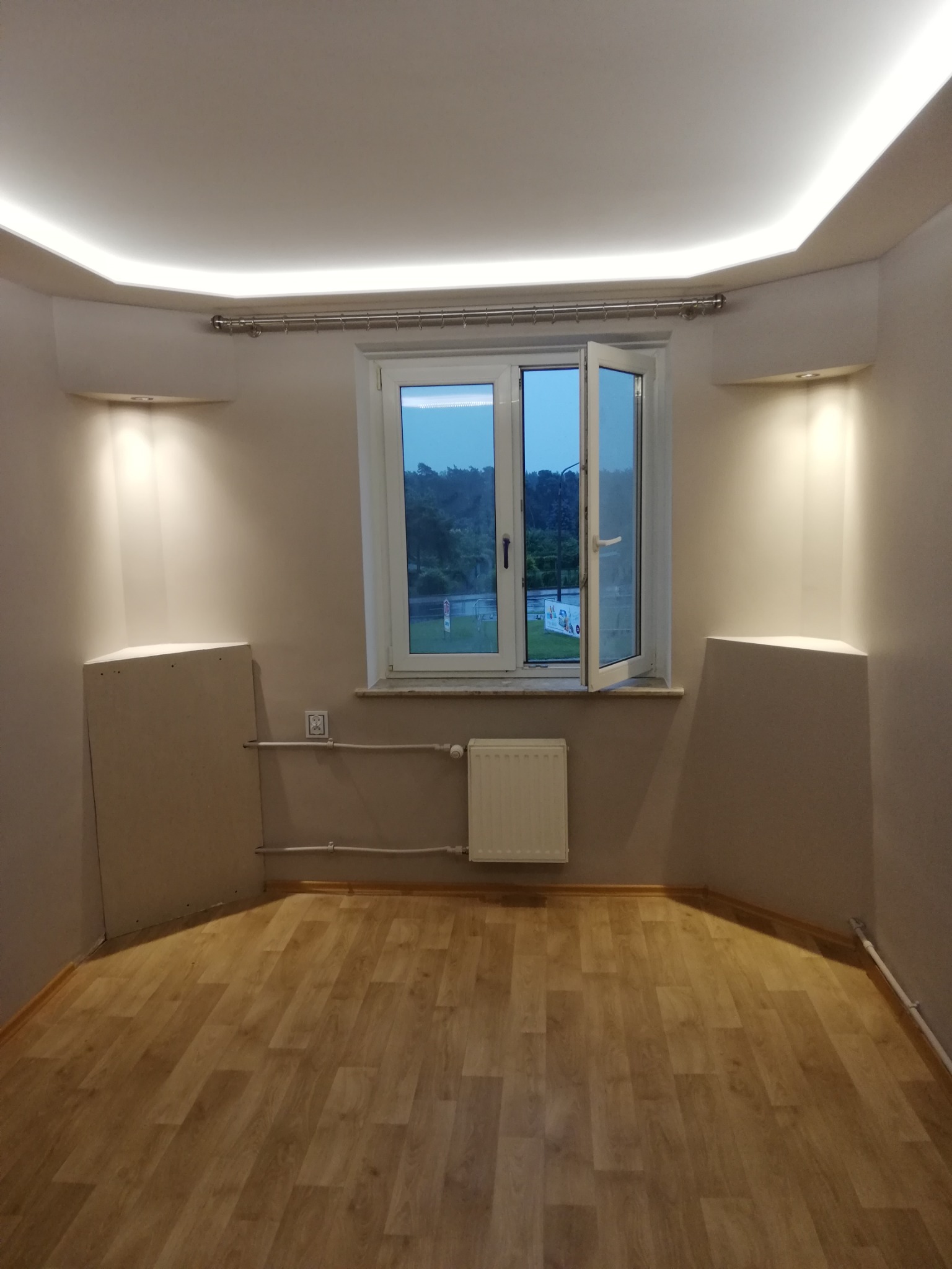 Pomalowane na jasny kolor wnętrze pokoju z drewnianą podłogą, otwartym oknem, grzejnikiem i dwoma surowymi, niedokończonymi zabudowami po bokach okna, oświetlonymi od góry punktowymi lampami.