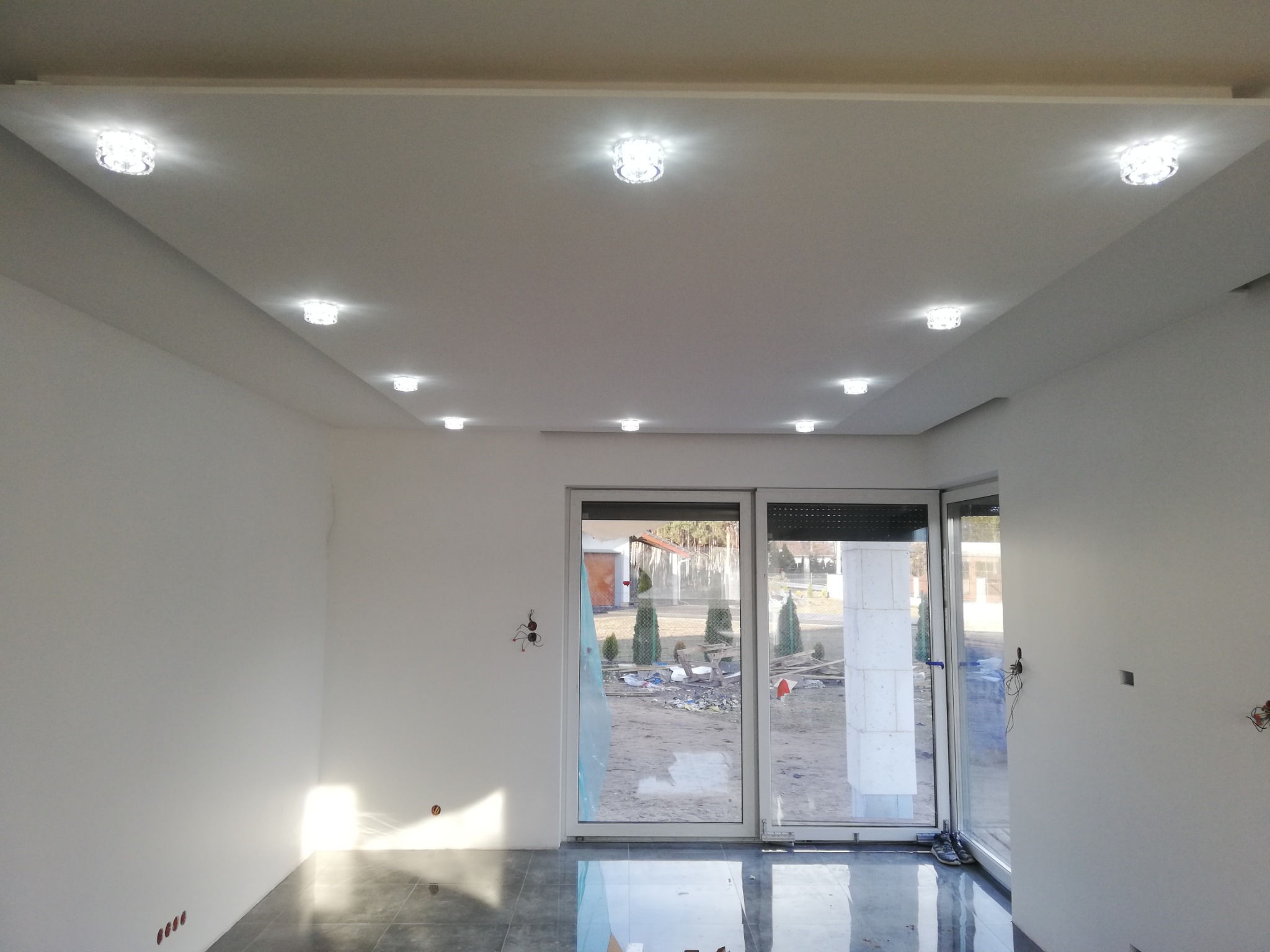 Jasne, przestronne wnętrze po remoncie, z błyszczącą szarą podłogą z płytek, białymi ścianami z widocznymi puszkami elektrycznymi i sufit podwieszany z wbudowanymi kryształowymi lampami. Przez duże...