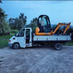 Biały samochód dostawczy z otwartymi drzwiami kierowcy, na którego pace stoi pomarańczowa mini koparka JCB 8014, teren trawiasto-żwirowy, pochmurne niebo.