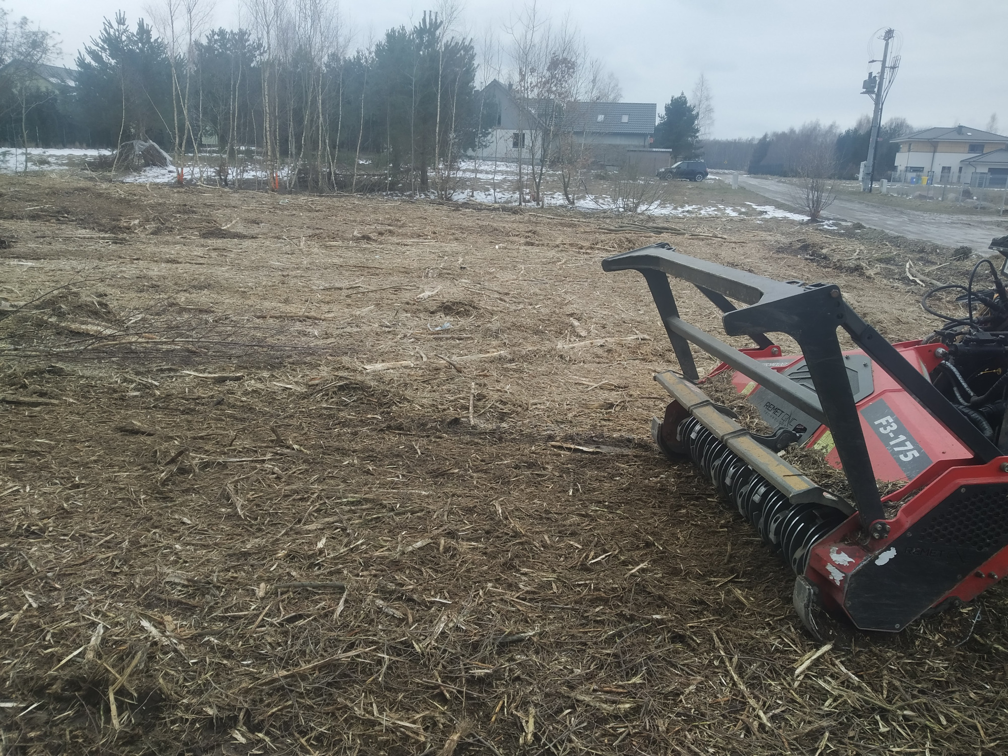 Teren po wycince samosiejek, pokryty zrębkami, z widoczną w tle zabudową jednorodzinną i fragmentem czerwonej frezarki do pni Remet CNC F3-175.