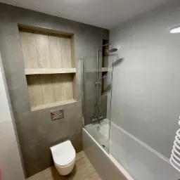 Nowoczesna łazienka z szarymi płytkami, drewnianą wnęką z półkami, podwieszaną toaletą i wanną z parawanem.