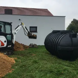 Czarny, żebrowany zbiornik na szambo betonowe z tworzywa sztucznego, przygotowany do zakopania, obok koparka Bobcat E19 z operatorem, wykop pod zbiornik oraz dom jednorodzinny w tle.