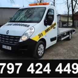 Biała laweta Renault z napisem 'Pomoc Drogowa 24h' i numerem telefonu na dole zdjęcia, zaparkowana na żwirowym podłożu przed budynkiem.