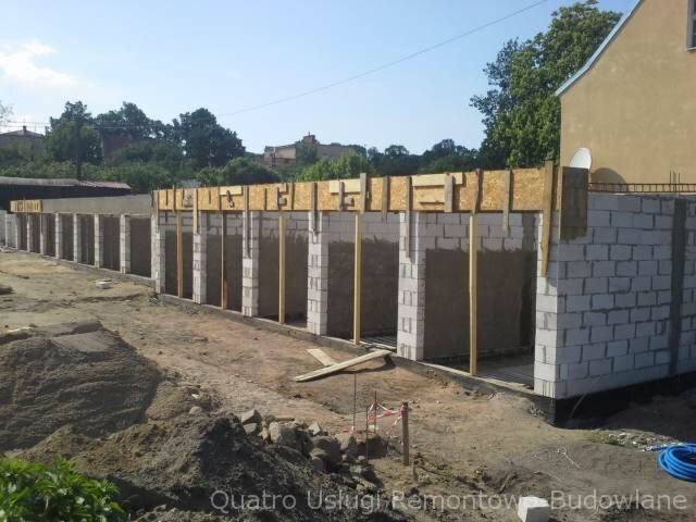 Wznoszenie ścian z bloczków betonowych z drewnianym szalunkiem, widok na plac budowy z hałdą ziemi i częściowo wykończonym budynkiem w tle.