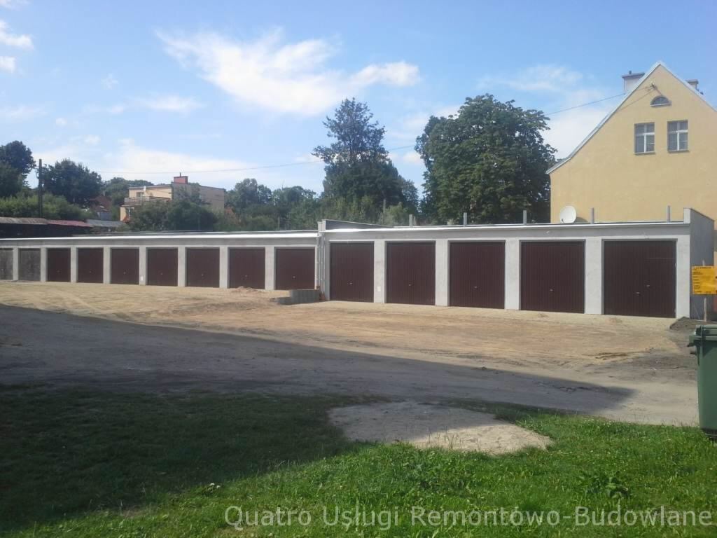 Rząd nowo wybudowanych garaży z brązowymi bramami segmentowymi, przy budynku mieszkalnym z satelitą na dachu, na tle błękitnego nieba.