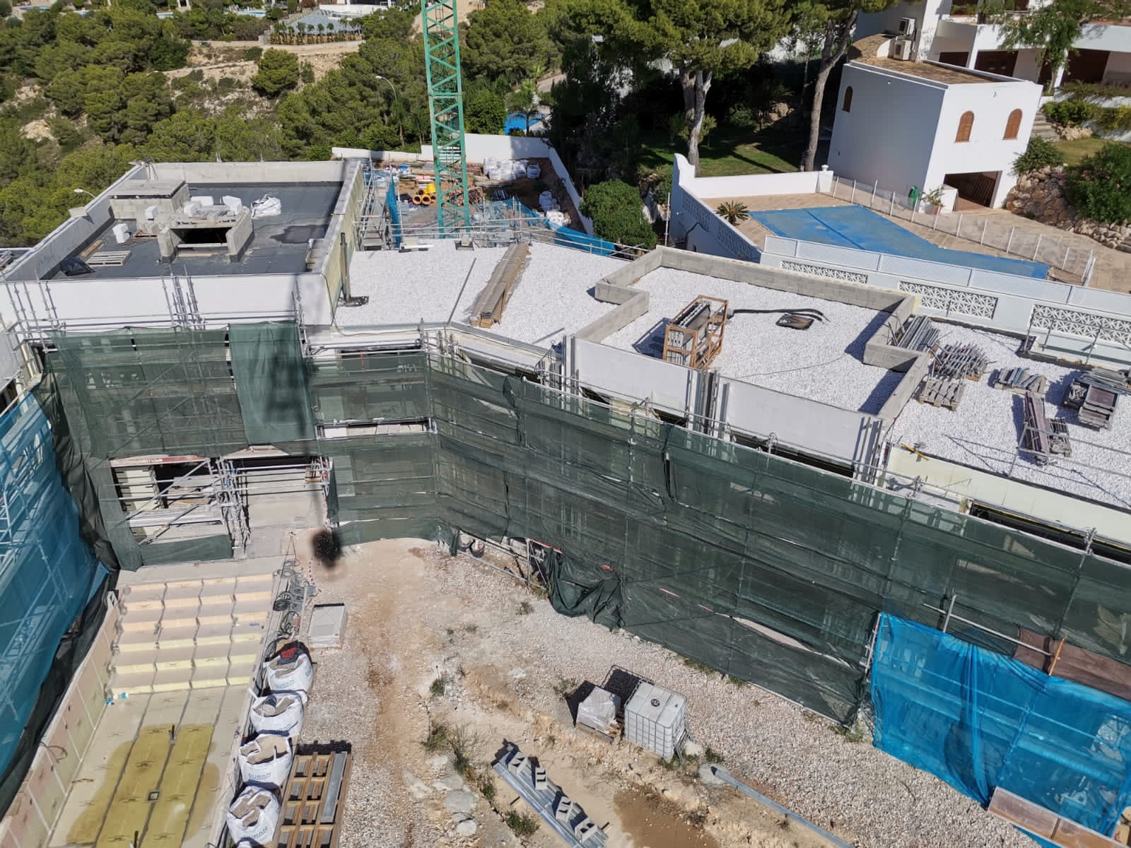 Wysoki kąt widokowy na budynek w trakcie budowy, osłonięty zieloną siatką rusztowań, z gruzem na dachu i dźwigiem w tle.