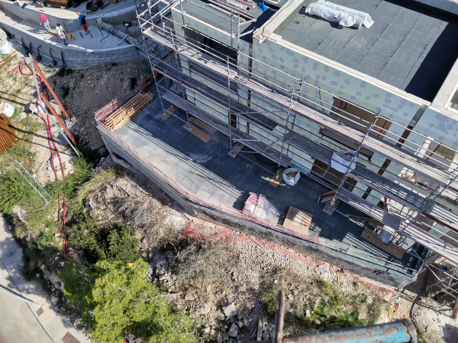 Widok z góry na budynek w trakcie budowy z rusztowaniami, widoczne elementy konstrukcyjne i materiały budowlane, teren wokół budynku z roślinnością.