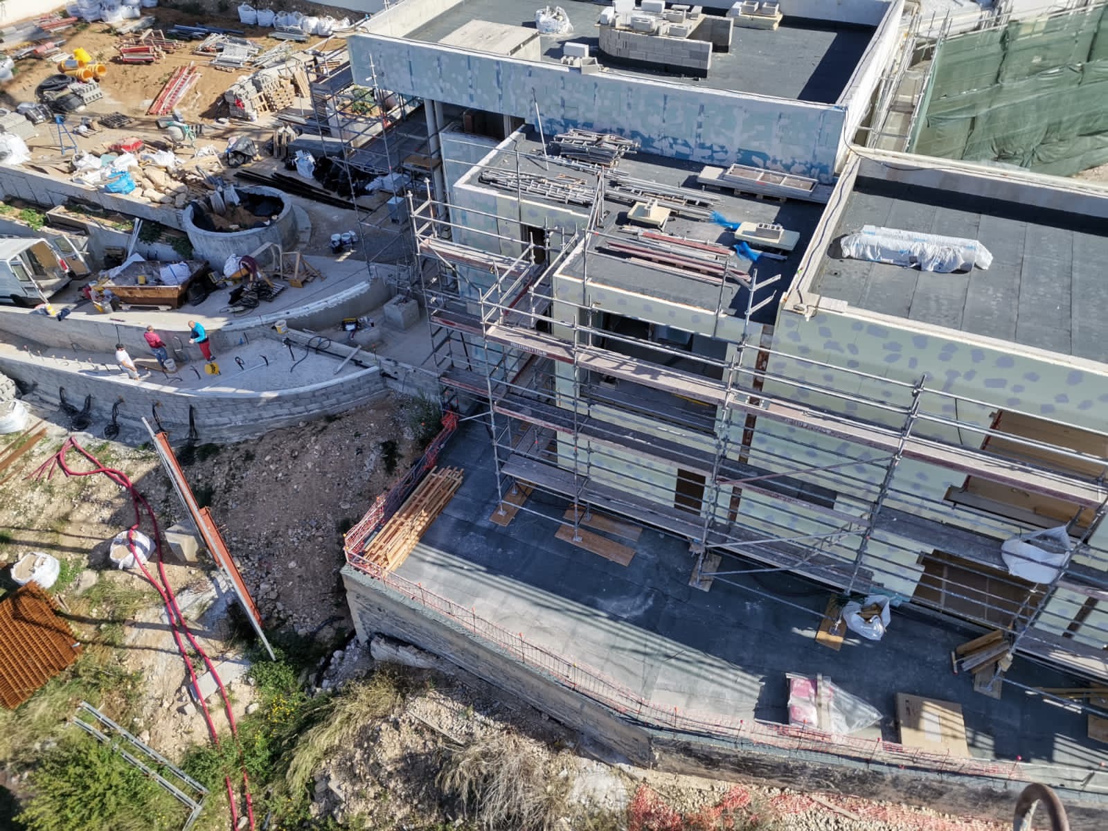 Budowa domu z widokiem z góry, widoczne rusztowania, ekipa budowlana pracuje przy betonowym murku oporowym, na dachu leżą materiały budowlane.