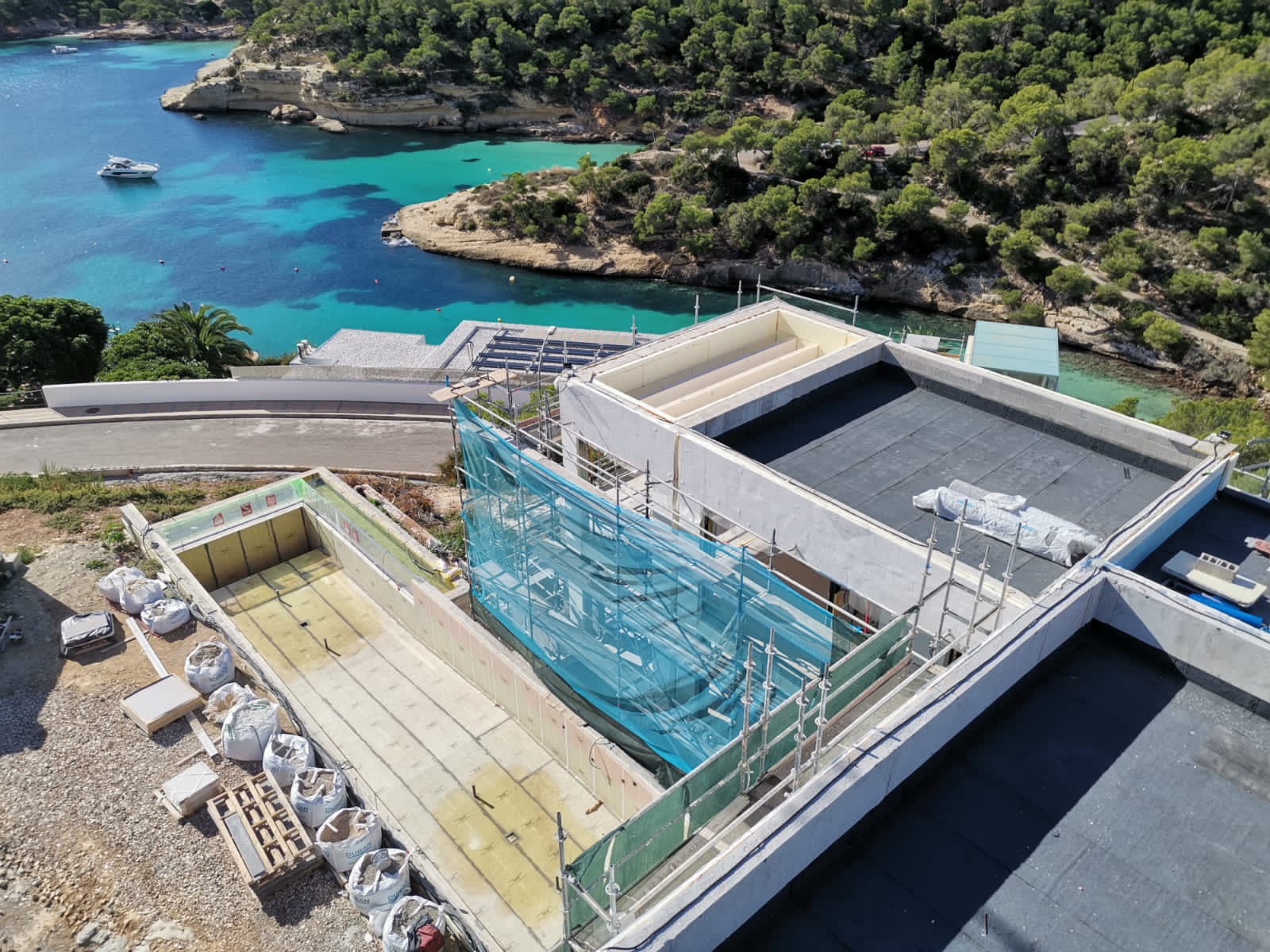 Widok z góry na budowę domu z basenem w otoczeniu turkusowej wody i zieleni, zabezpieczony rusztowaniami i siatką.