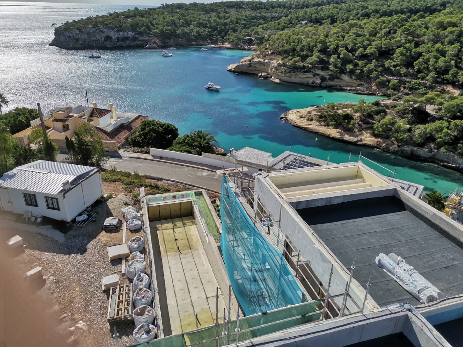 Widok z góry na budowę domu z widokiem na turkusową zatokę, widoczne niezadaszone, betonowe ściany, materiały budowlane w workach i zwinięta papa na dachu.
