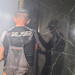 Remonty i wykończenia - Pracownik w kombinezonie montuje płytki imitujące czarny marmur w łazience, widoczne krzyżyki dystansowe i wystające rury instalacji wodnej.