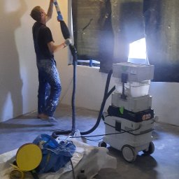 Remonty i wykończenia - Pracownik w kombinezonie szlifuje sufit za pomocą szlifierki z odkurzaczem przemysłowym, okno zabezpieczone folią.