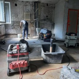 Remonty i wykończenia - Wnętrze w trakcie prac przygotowawczych do remontu: widoczne dwa odkurzacze przemysłowe, kuweta, pracownicy w tle, gołe ściany i sufit w stanie surowym.
