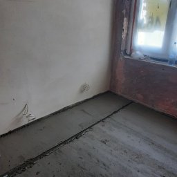 Remonty i wykończenia - Surowy, szary beton na podłodze w trakcie wylewania, widoczne listwy dylatacyjne, ściany w stanie surowym, okno zabezpieczone folią malarską.