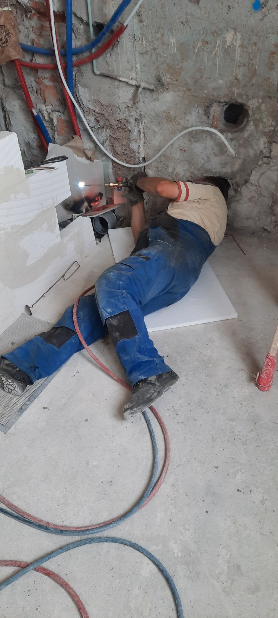 Instalator w niebieskim kombinezonie spawa rurę w surowym wnętrzu, widoczne przewody instalacji wodnej i fragmenty muru.