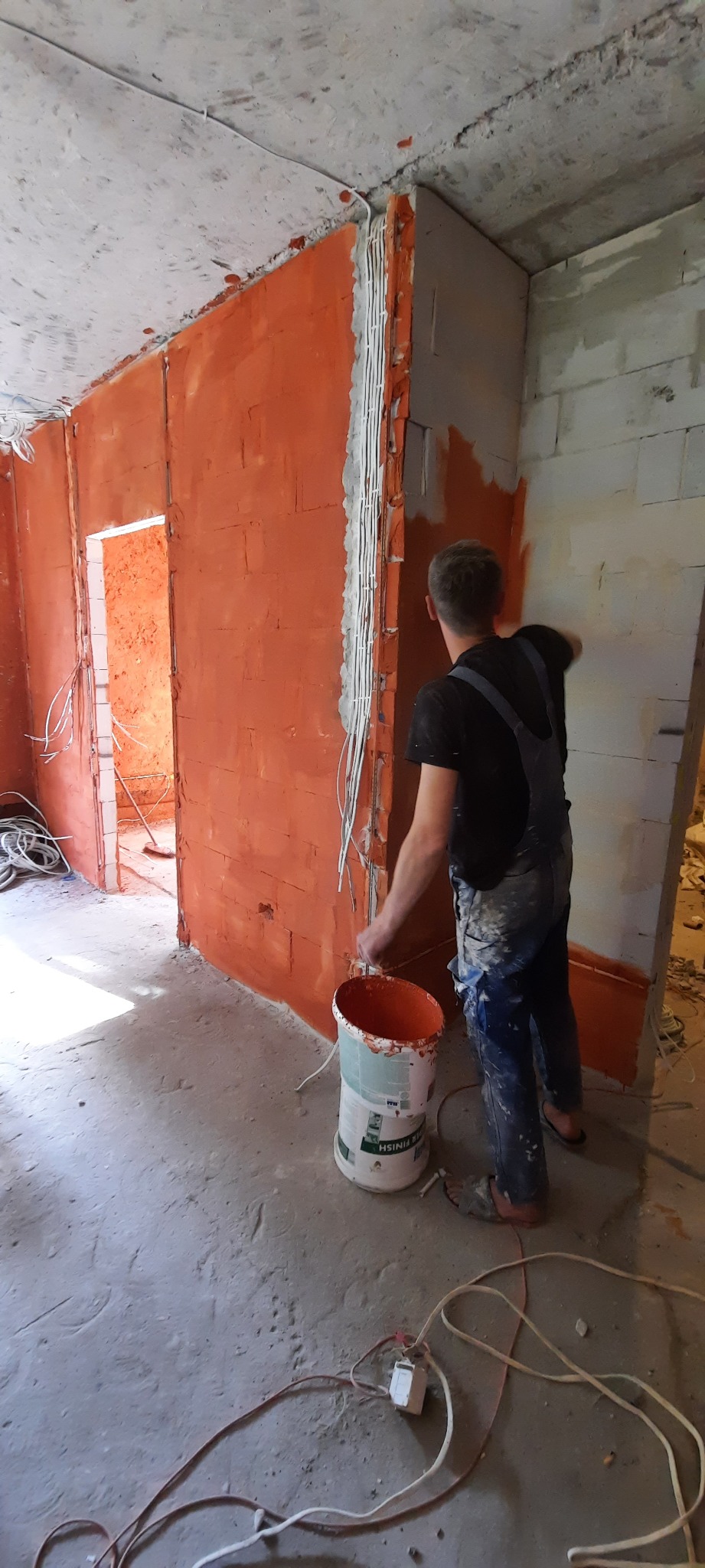 Mężczyzna w kombinezonie malarskim nakłada pomarańczową farbę na ścianę z cegły w surowym wnętrzu, widoczne przewody elektryczne i wiadro z farbą.