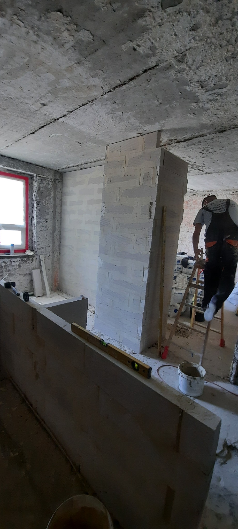 Wnętrze w trakcie prac murarskich: wznoszenie ścianki działowej z bloczków, widoczny pracownik na drabinie, surowy betonowy strop, okno z czerwoną ramą, narzędzia i materiały budowlane.