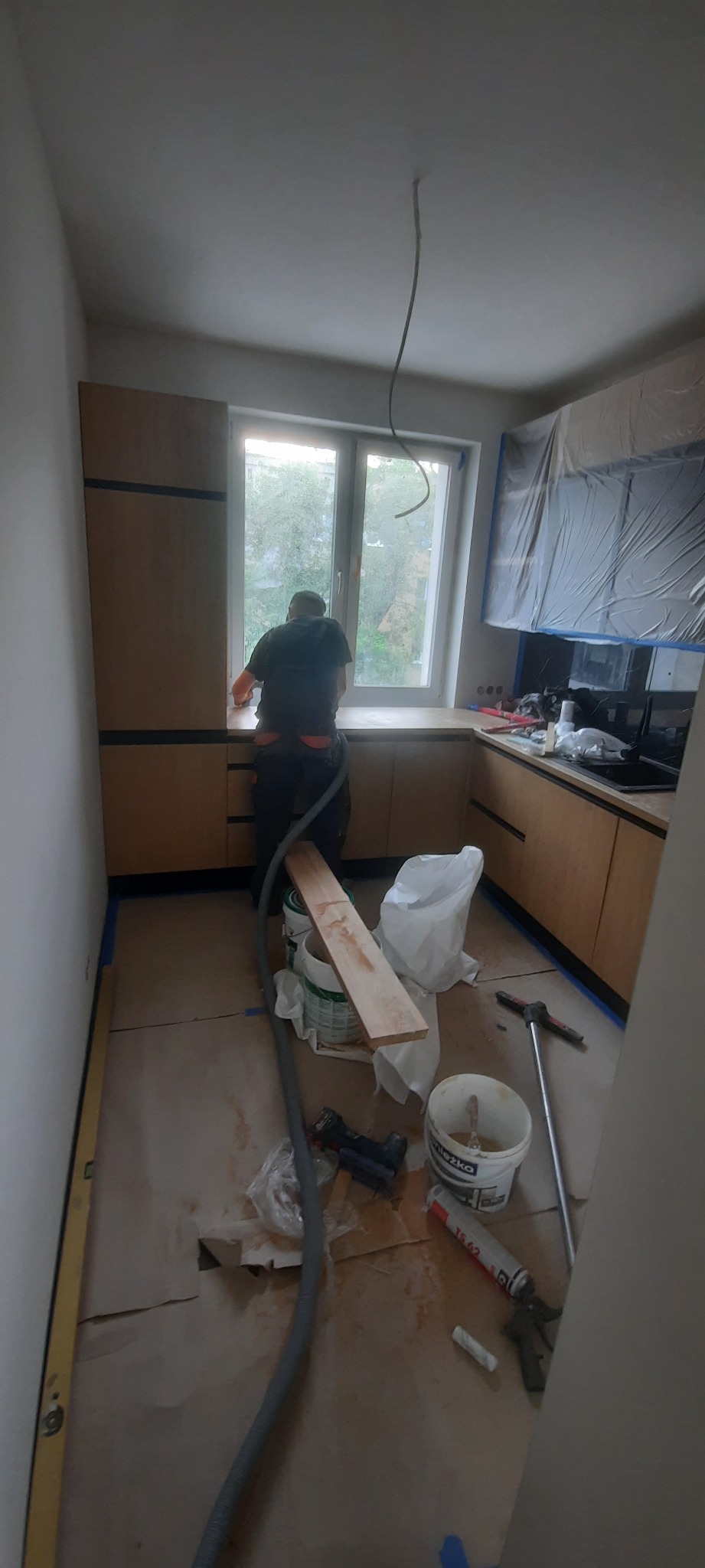 Pracownik w trakcie montażu blatu kuchennego przy oknie, widoczne narzędzia, poziomica, wiadro z klejem, zakurzona podłoga zabezpieczona kartonem, osłonięte folią szafki wiszące.