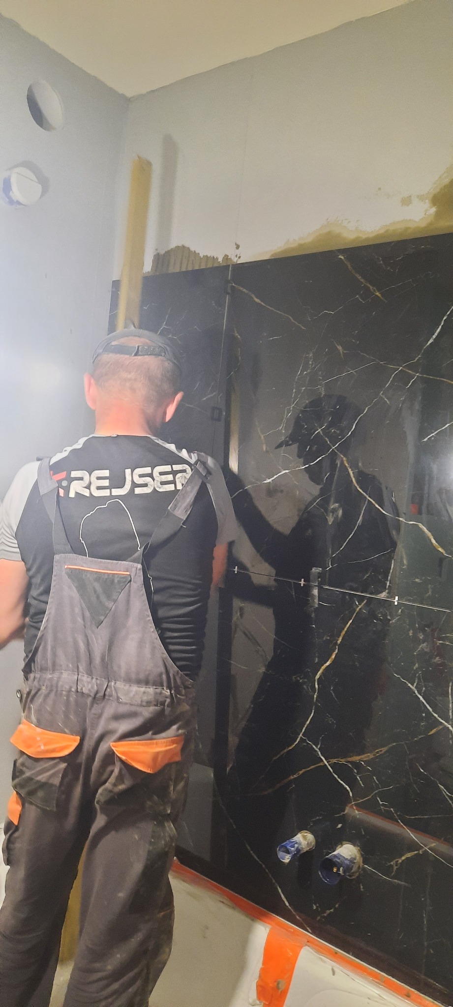 Pracownik w kombinezonie montuje płytki imitujące czarny marmur w łazience, widoczne krzyżyki dystansowe i wystające rury instalacji wodnej.