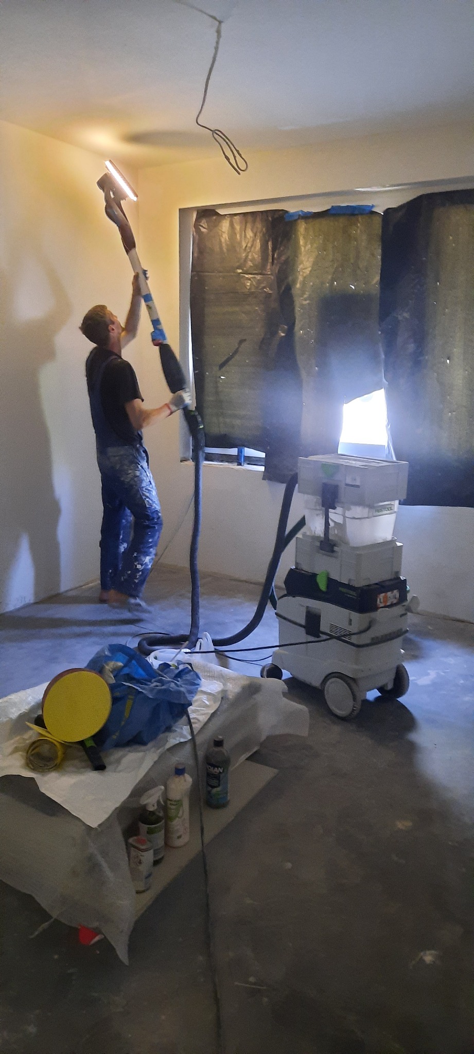 Pracownik w kombinezonie szlifuje sufit za pomocą szlifierki z odkurzaczem przemysłowym, okno zabezpieczone folią.