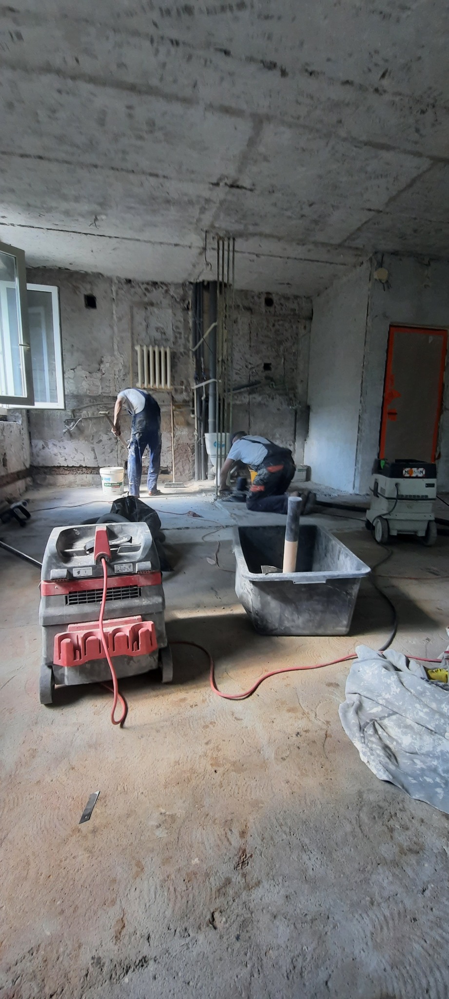 Wnętrze w trakcie prac przygotowawczych do remontu: widoczne dwa odkurzacze przemysłowe, kuweta, pracownicy w tle, gołe ściany i sufit w stanie surowym.